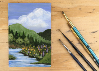 An acrylic painting of the Wasatch Mountains on a wooden table with paintbrushes next to it.