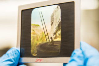 An anthropologist holds a lantern slide of a Native American man climbing a wooden ladder out of a kiva.
