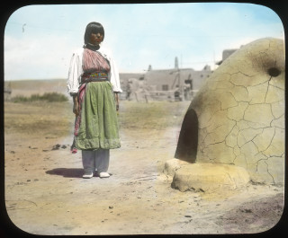 A lantern slide of a Native American woman standing next to a clay oven.