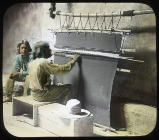 A Native American man weaves a rug on a loom.