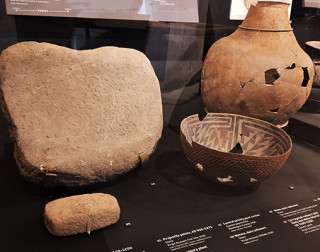 A mano, metate, cracked bowl, and jug in a display case