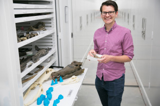 A scientist smiles for the camera while standing next to a cabinet of fossils. 