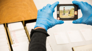 An anthropologist holds a lantern slide featuring a Native American woman standing next to a clay oven.