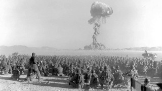 A distance mushroom cloud from a test explosion is observed by a large group of soldiers. 