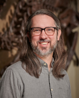 Dr. David Evans smiles standing in front of a dinosaur skeleton.