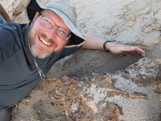 Dr. Andrew Farke smiles in front of a quarry of fossils embedded into rock.