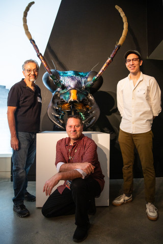 Three men pose for a photo next to a large model of a tiger beetle head. 