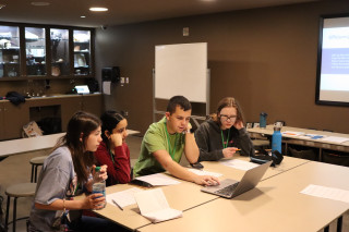 4 teens sit at a table looking at a laptop
