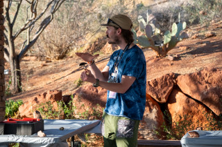 An educator in a tie dye shirt and hat holds up a mushroom in front of a desert background