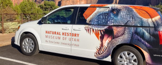 A white outreach van with a carnivorous dinosaur is parked in front of scenic red rock cliffs