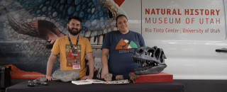 Educators Nichelle and Jacob smile in front of the NHMU Mobile Museum with a table of fossils in front of them.