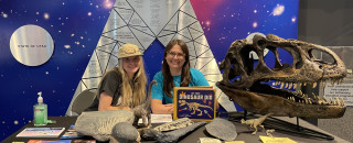 Layne Sermersheim (Statewide Outreach Coordinator) and Katie Vitti (Wasatch Front Outreach Coordinator) sitting at their outreach table which features a variety of dinosaur fossils, like an allosaurus skull.