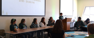 1 teen stands at a podium while 5 sit at a table in front of a room