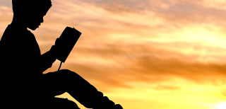 A silhouette of a boy reading a book against a sunset. 