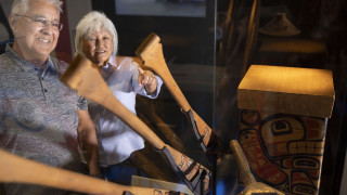 An older couple view indigenous artwork, including paddles, through a glass case. 