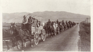 A historic black and white photo of a wagon train following a road.