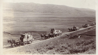 A historic black and white photo of a wagon train following a road.