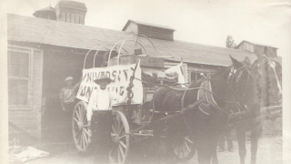 A historic black and white photo of a wagon outside a building.