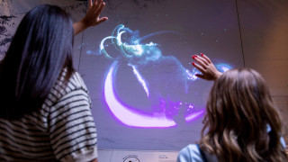 A mother and daughter wave at a projection in an exhibit revealing a mammoth.