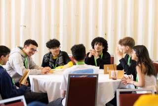 7 young adults and teens gathered around a table talking with each other.