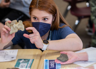 An NHMU educator wearing a face masks shows a fossil to students at a table