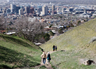 4 people are seen walking down a trail through a green meadow with downtown Salt Lake in the background. 