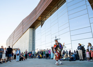 A Native American man in traditional regalia dances before a crowd in front of a large building