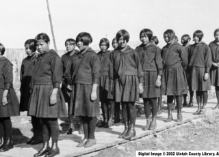 A black and white photo of young Ute girls wearing school uniforms standing in line. 