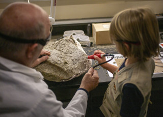 A young person with the assistance of an adult uses an air drill on a piece of rock 