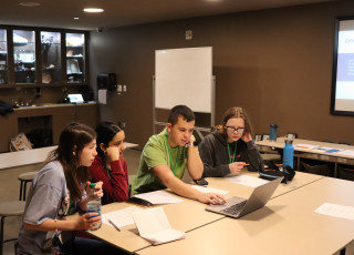 4 teens sit at a table and look at a laptop