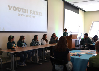 A teen stands at a podium while 4 teens sit at a table in front of a room
