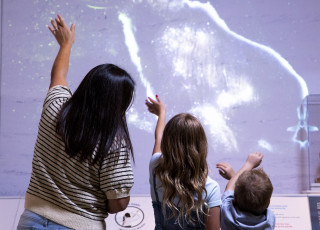 an adult and two children hold up their arms before a digital display of a mammoth