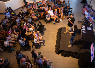 A picture of a large crowd gathered around 2 people speaking on a small stage